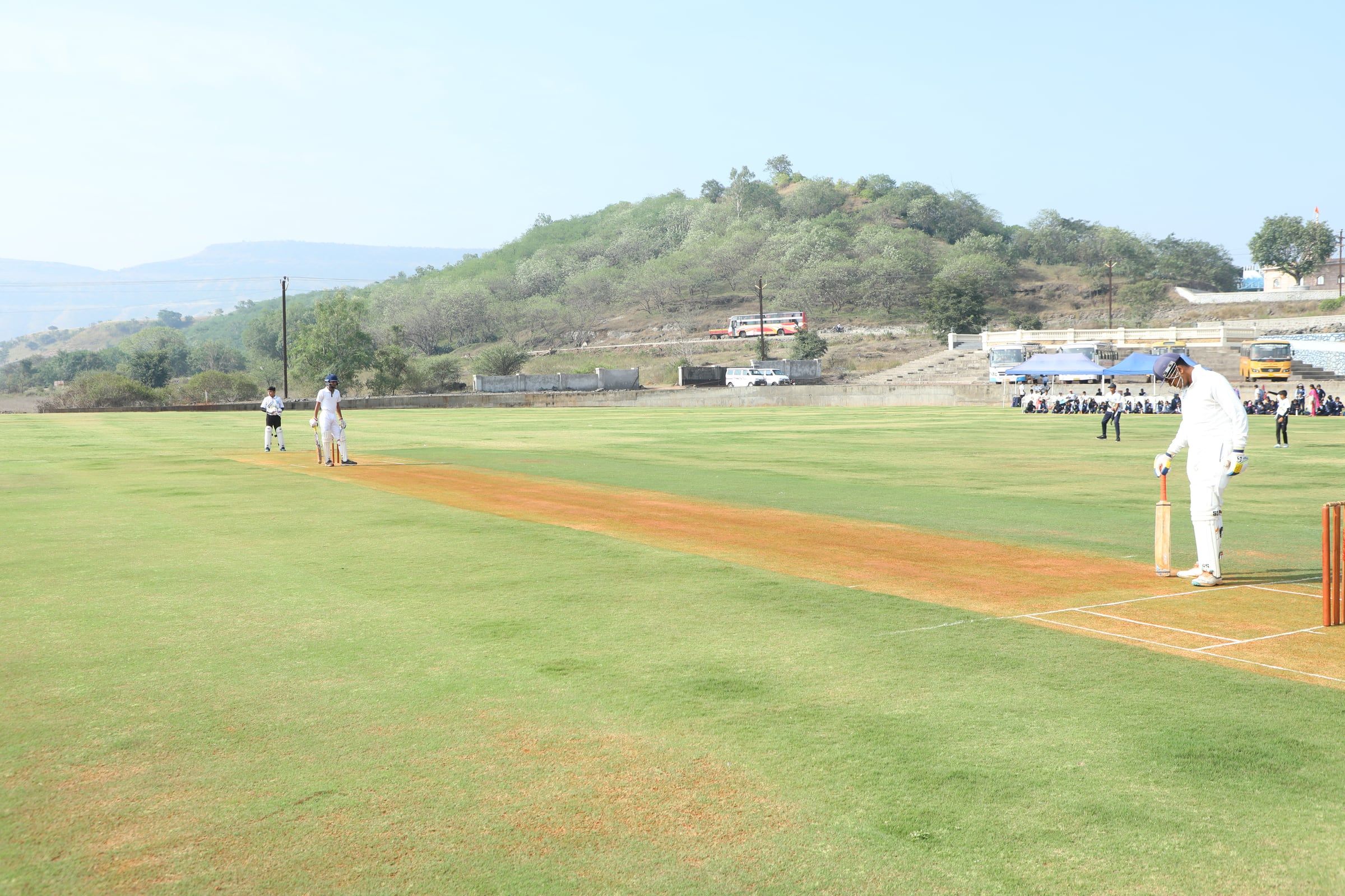 A cricket match on a green ground with a hill behind it