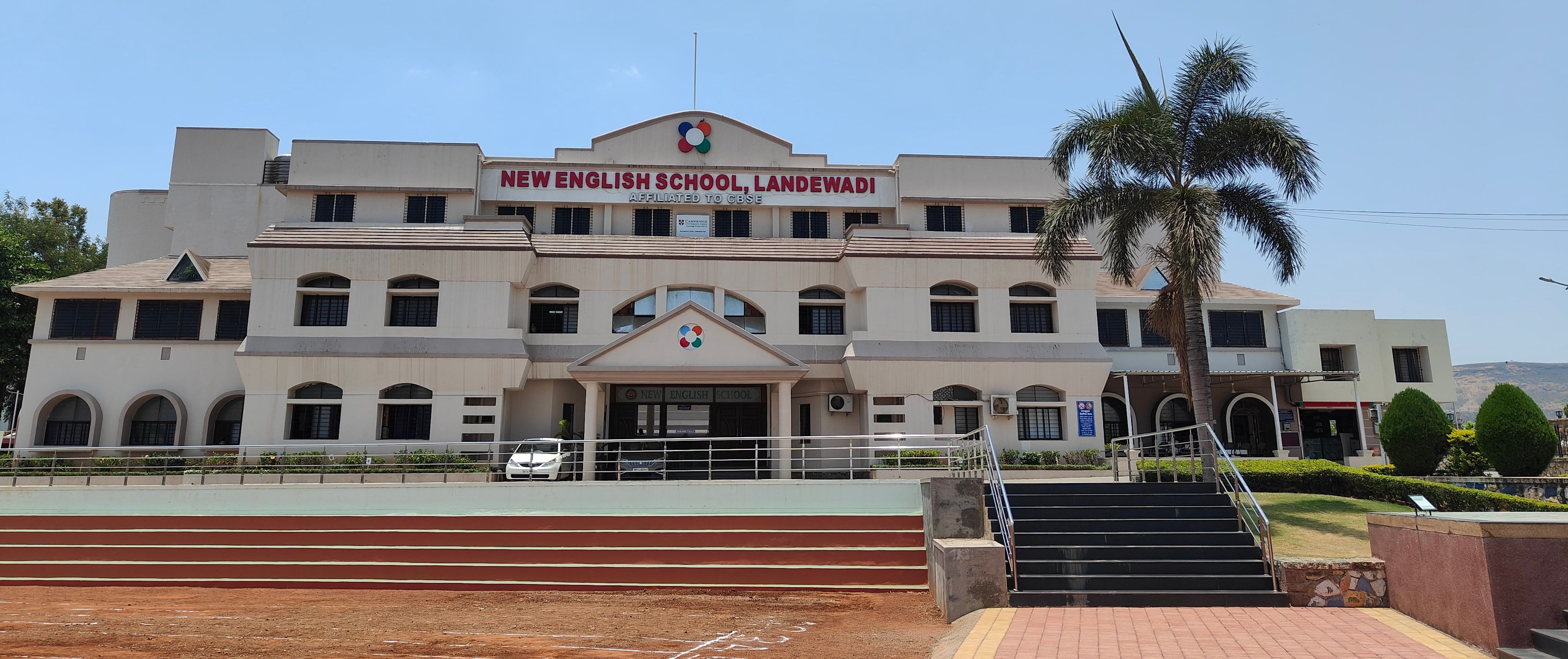 New English School, Landewadi: a three-storey cream building with arched windows, a palm tree and a red running track in front