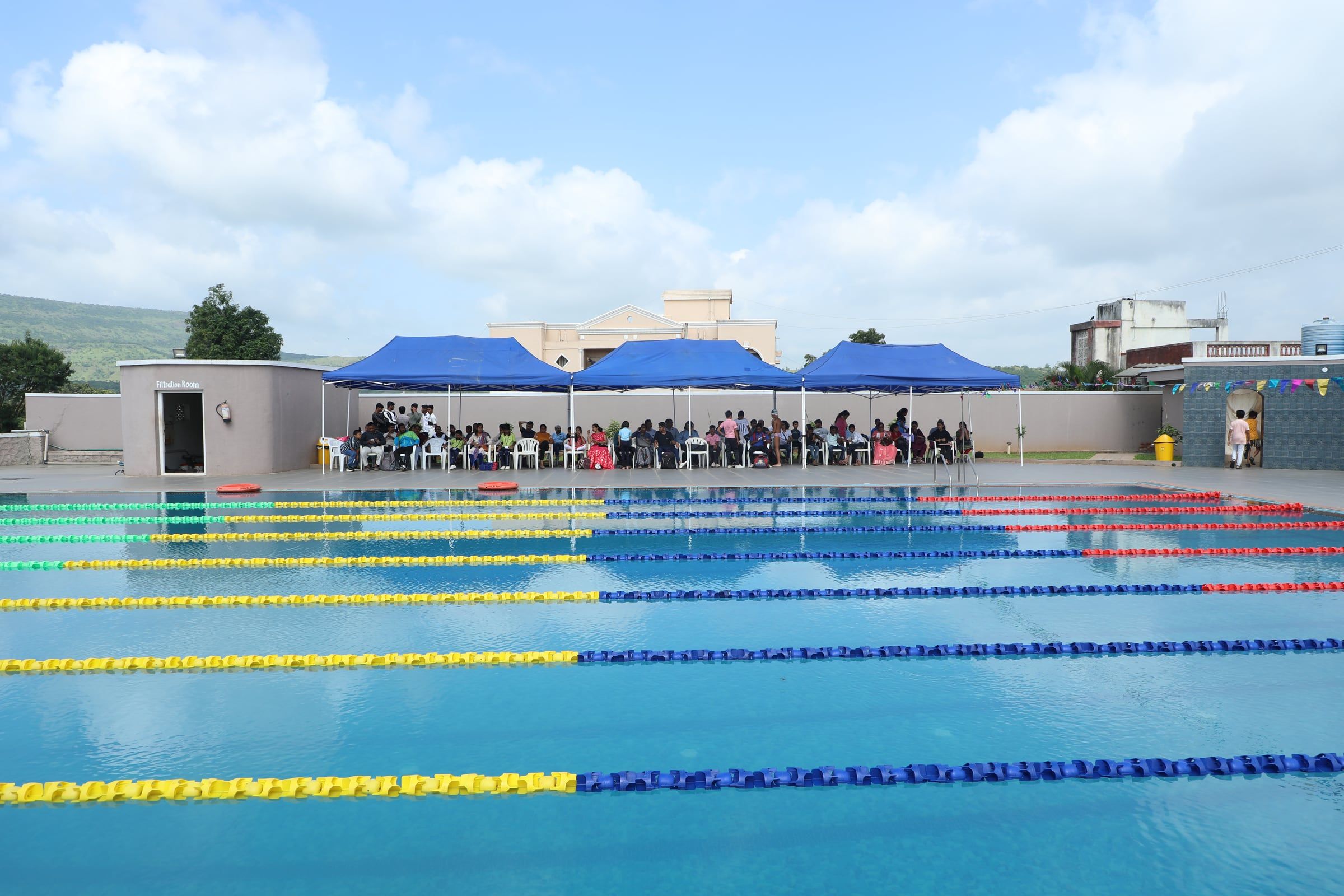 An outdoor swimming pool with lane ropes and spectators under blue canopies