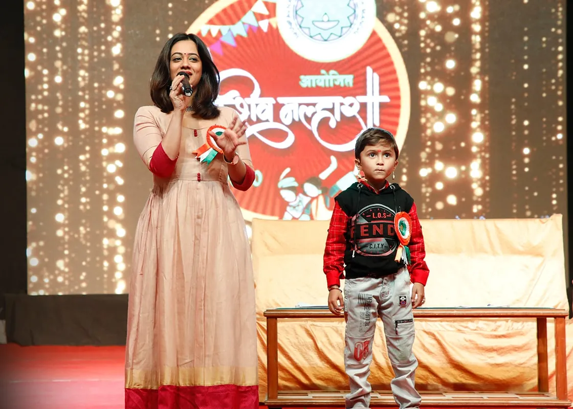 A presenter with a microphone beside a young student on the Shiv Malhar stage